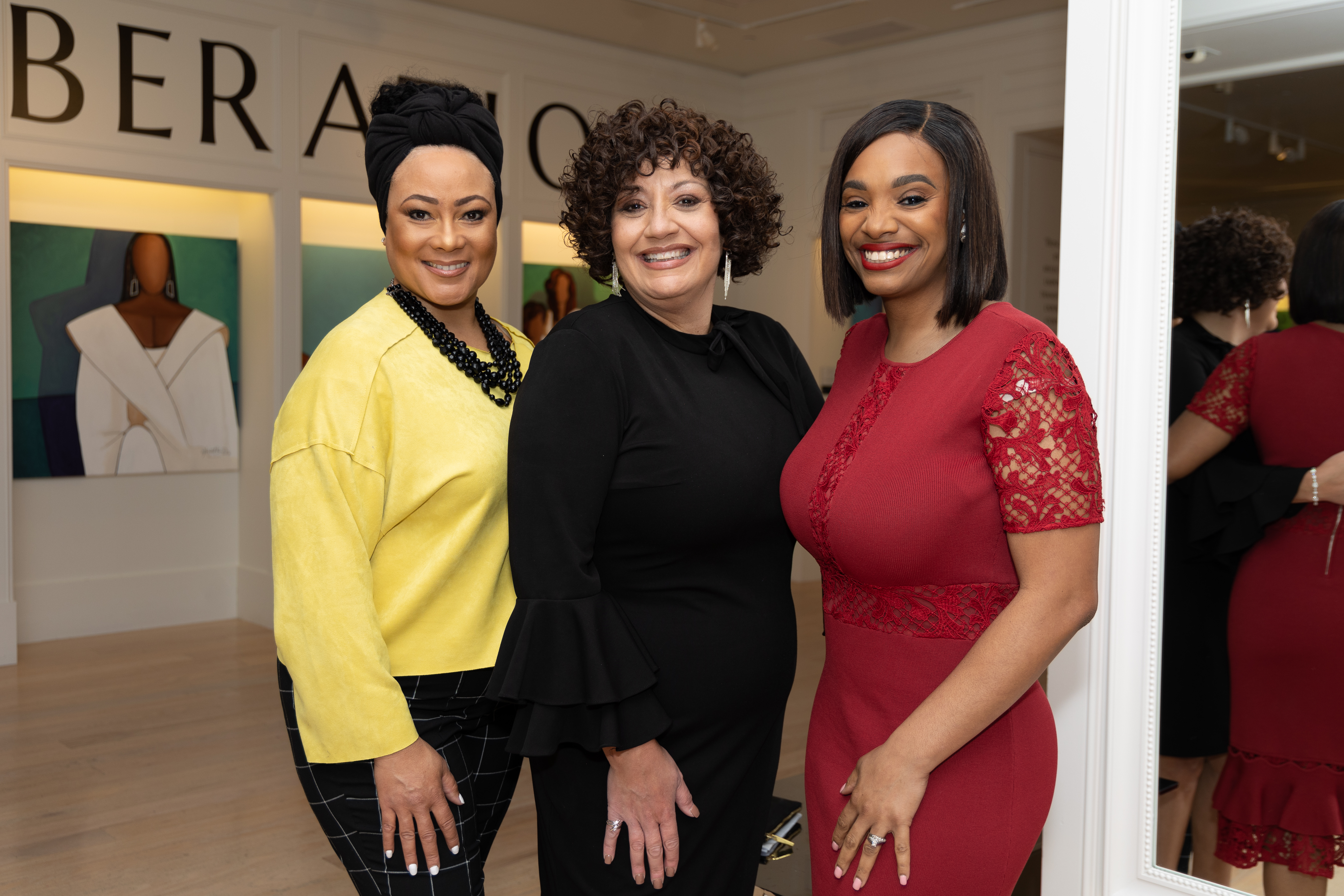 Three black women standing together: one in a black head wrap and yellow top, one with curly hair in a black dress, and one with a straight shoulder-length bob in a red dress.