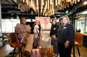 1. Five women (moderator Director Gregory left, panelists right) smile in their seats towards camera. Room is brightly lit and warm. Panelist Rep. Tarra Simmons holds her puppy.