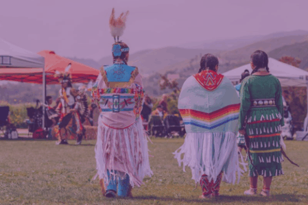 Three native women in regalia walk with backs turned to the camera